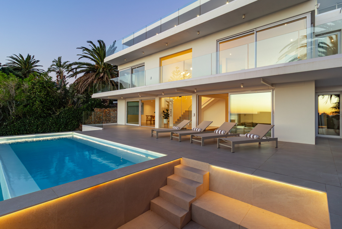 A luxury villa situated in Bakoven, next to Camps Bay in the city of Cape Town, South Africa. It shows the back courtyard house view showing double storey house with glass balcony, sliding doors, tiled deck with lounge chairs, and part of a ground floor level blue rectangle pool with stairs next to it leading to lower level part of the property.