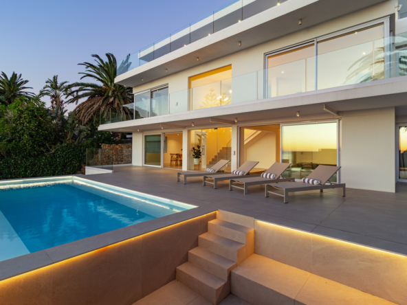 A luxury villa situated in Bakoven, next to Camps Bay in the city of Cape Town, South Africa. It shows the back courtyard house view showing double storey house with glass balcony, sliding doors, tiled deck with lounge chairs, and part of a ground floor level blue rectangle pool with stairs next to it leading to lower level part of the property.