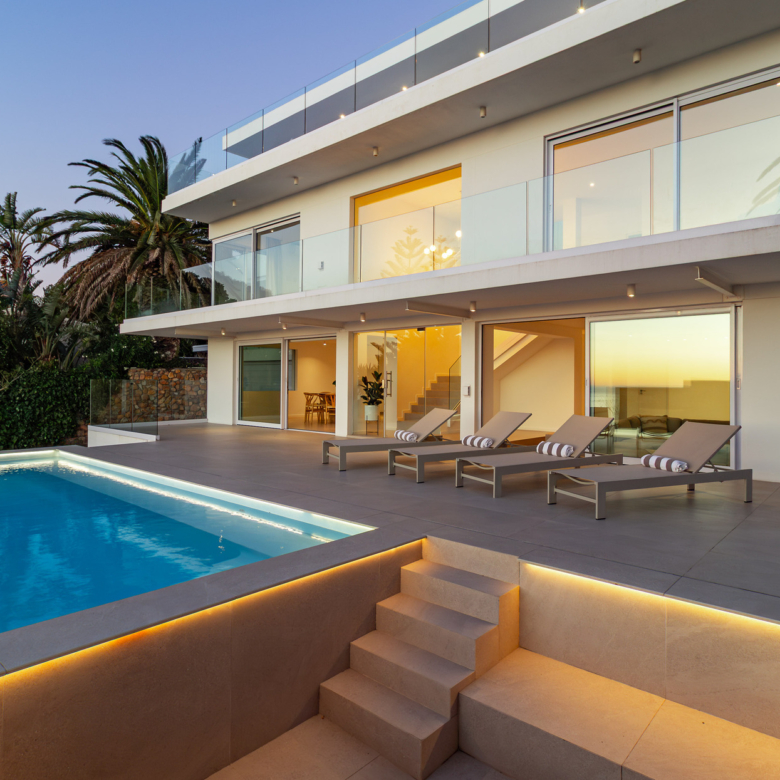 A luxury villa situated in Bakoven, next to Camps Bay in the city of Cape Town, South Africa. It shows the back courtyard house view showing double storey house with glass balcony, sliding doors, tiled deck with lounge chairs, and part of a ground floor level blue rectangle pool with stairs next to it leading to lower level part of the property.