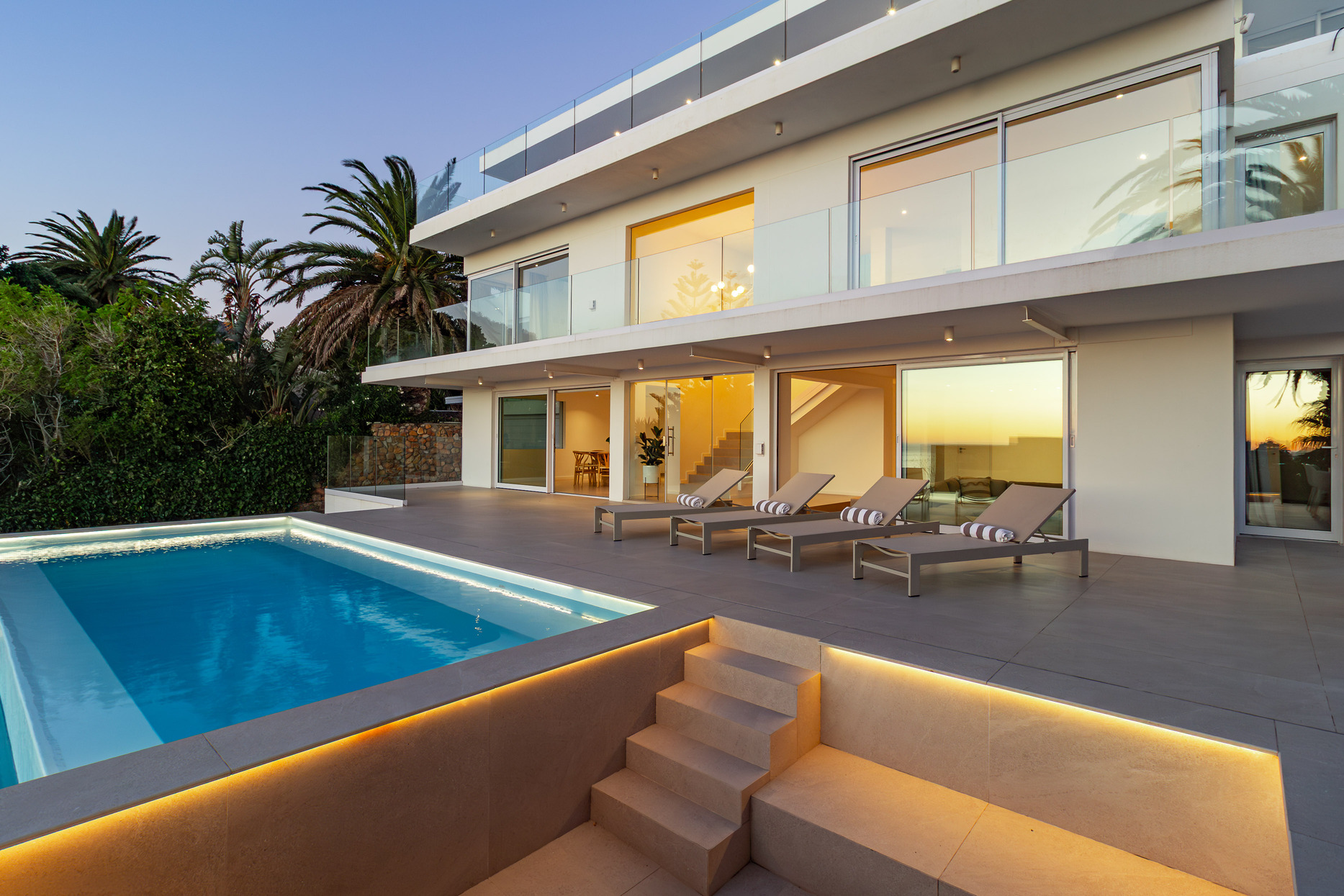 A luxury villa situated in Bakoven, next to Camps Bay in the city of Cape Town, South Africa. It shows the back courtyard house view showing double storey house with glass balcony, sliding doors, tiled deck with lounge chairs, and part of a ground floor level blue rectangle pool with stairs next to it leading to lower level part of the property.
