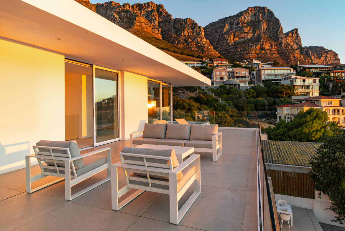 23odk luxury villa balcony with outdoor lounge furniture showing view of the twelve apostles mountain and few houses at early sunset in Bakoven next to Camps Bay, a coastal town in Cape Town, South Africa.