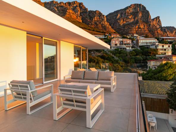23odk luxury villa balcony with outdoor lounge furniture showing view of the twelve apostles mountain and few houses at early sunset in Bakoven next to Camps Bay, a coastal town in Cape Town, South Africa.