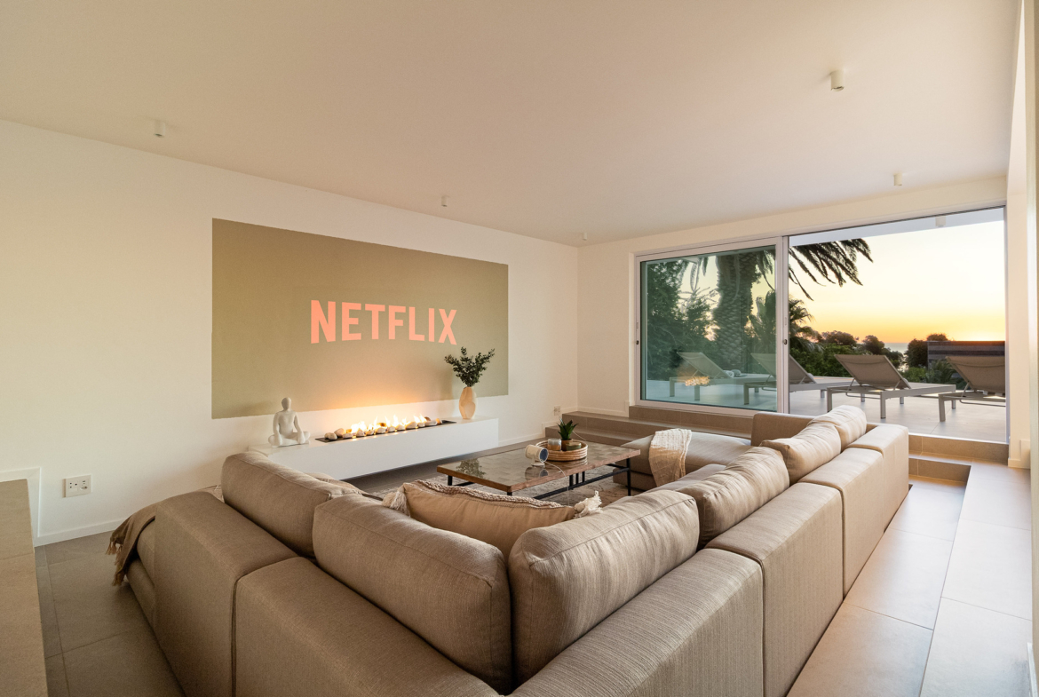 A luxury villa vacation rental situated in Bakoven, next to Camps Bay in the city of Cape Town, South Africa. This photo shows a sunken lounge with a snugly fit large comfortable L-shaped modular sofa, coffee table and cinema wall projection image. It has large open sliding doors showing part of the pool deck area, lounge chairs, palm trees and an early sunset view.