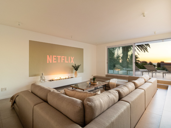 A luxury villa vacation rental situated in Bakoven, next to Camps Bay in the city of Cape Town, South Africa. This photo shows a sunken lounge with a snugly fit large comfortable L-shaped modular sofa, coffee table and cinema wall projection image. It has large open sliding doors showing part of the pool deck area, lounge chairs, palm trees and an early sunset view.