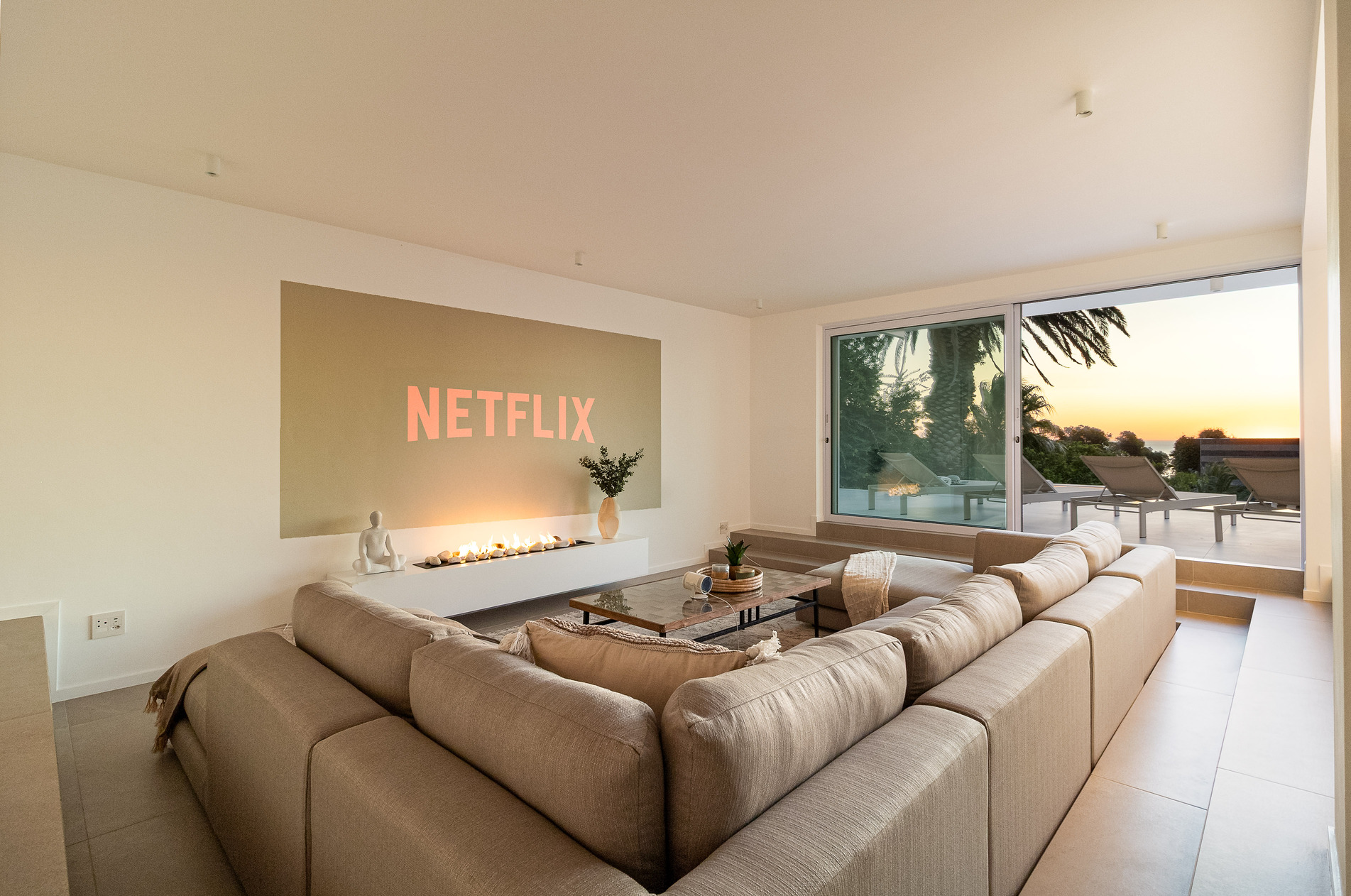 A luxury villa vacation rental situated in Bakoven, next to Camps Bay in the city of Cape Town, South Africa. This photo shows a sunken lounge with a snugly fit large comfortable L-shaped modular sofa, coffee table and cinema wall projection image. It has large open sliding doors showing part of the pool deck area, lounge chairs, palm trees and an early sunset view.